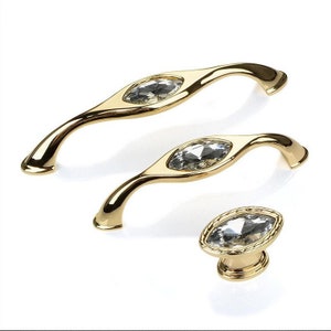 Pode incluir: Três botões de armário dourados com detalhes em cristal transparente. Os botões têm a forma de uma lágrima com uma pega curva.