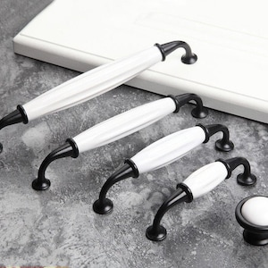 以下が含まれることがあります： 黒色の金属製の土台に、白いセラミック製のキャビネットノブが4つあります。ノブはサイズが異なり、大きい順に並んでいます。