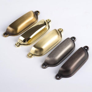 Peut inclure: Quatre poignées de tiroir en différentes finitions : laiton antique, laiton poli, nickel brossé et bronze huilé. Chaque poignée a une forme rectangulaire incurvée avec des bords arrondis.