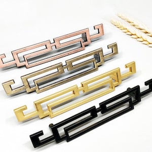 Könnte beinhalten: Vier dekorative Schrankgriffe in verschiedenen Ausführungen: Roségold, Silber, Gold und Schwarz. Jeder Griff hat ein geometrisches Design mit quadratischer Form.