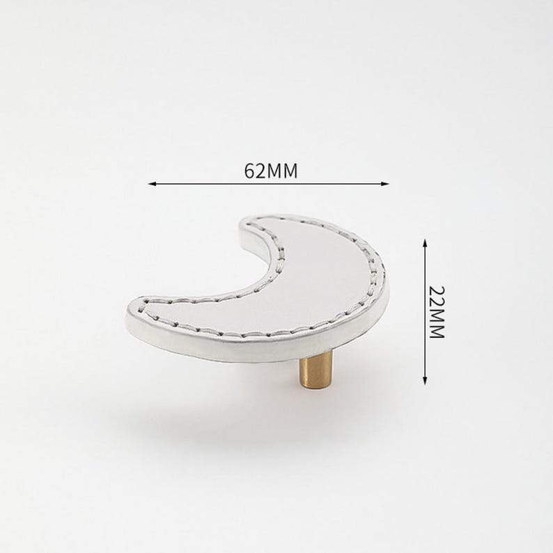 May include: A white crescent moon-shaped cabinet knob with a stitched edge and a brass base. The knob measures 62 mm wide and 22 mm high.