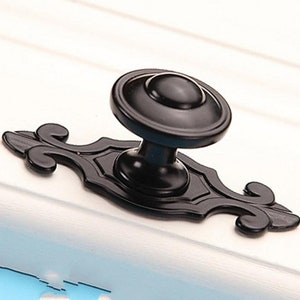 Może przedstawiać: Czarny gałka do szafki z ozdobną, zdobioną podstawą. Gałka jest okrągła i ma gładkie, polerowane wykończenie.