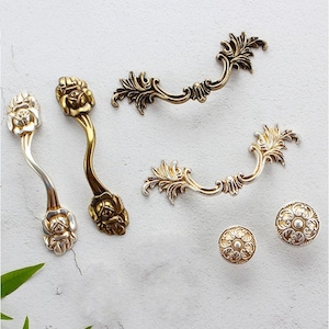 Könnte beinhalten: Sortiment an dekorativer Schrankbeschläge in Gold- und Silberfarben. Die Beschläge umfassen zwei verzierte Schubladenzüge mit Blumendesigns, zwei verzierte Schubladenzüge mit Blattmustern und zwei runde Knöpfe mit Blumendesigns.