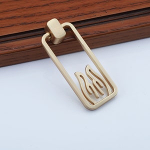 May include: Gold-toned metal cabinet pull with a rectangular shape and a cutout design of stylized flames.
