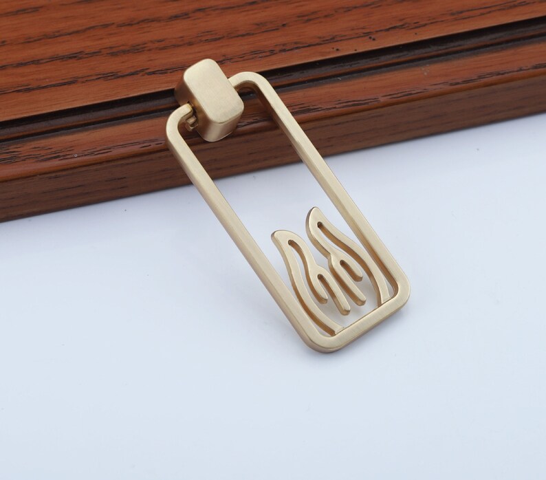 May include: Gold-toned metal cabinet pull with a rectangular shape and a cutout design of stylized flames.