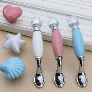 May include: Four ceramic cabinet knobs in white, pink, and blue with a silver metal base. The knobs are shaped like a seashell, a starfish, a heart, and an oval.