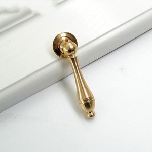 Könnte beinhalten: Ein goldfarbener Schrankgriff mit abgerundeter Oberseite und einem langen, schlanken Griff. Der Griff hat eine leichte Biegung und eine glatte, polierte Oberfläche.