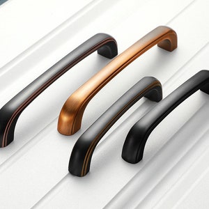 May include: Four cabinet pulls in two finishes: black and copper. The pulls are all the same shape, with a curved top and bottom and a straight middle section.
