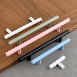 Könnte beinhalten: Fünf Schrankgriffe in verschiedenen Farben: Weiß, Hellgrün, Rosa, Schwarz und Hellblau. Die Griffe haben alle die gleiche Größe und Form mit einem einfachen, modernen Design.