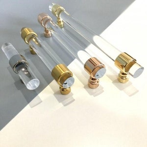 May include: Four clear acrylic cabinet pulls with different metal finishes: gold, rose gold, and silver. The pulls are all the same size and shape, with a cylindrical body and a rounded end.