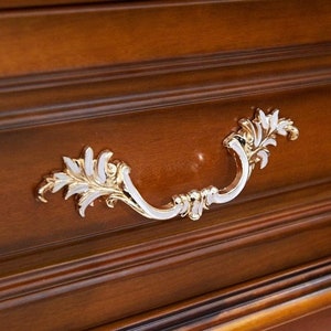 May include: An ornate drawer pull with a leaf design, finished in white and gold. The pull is mounted on a wooden drawer front.