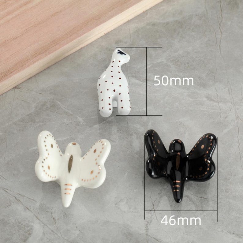 May include: Three ceramic decorative pieces: a giraffe-shaped figurine (50mm tall) with brown spots, and two butterfly-shaped figurines (46mm wide), one white with gold accents, and one black with gold accents. The pieces are on a gray surface.