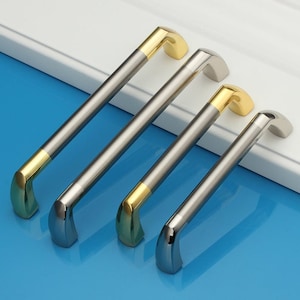 Op de afbeelding: Vier kastgrepen met een geborsteld nikkel afwerking en een gouden accent. De grepen zijn gerangschikt in een rij van groot naar klein.