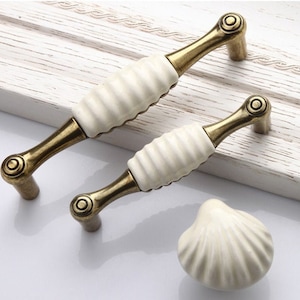 May include: Two white ceramic cabinet knobs with a ribbed design and antique brass hardware. A matching white ceramic shell-shaped knob is also shown.