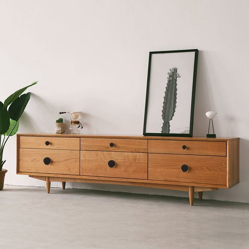 May include: A long, wooden dresser with six drawers and black round knobs. The dresser is sitting on a light grey floor in front of a white wall. A framed print of a cactus is hanging on the wall above the dresser.