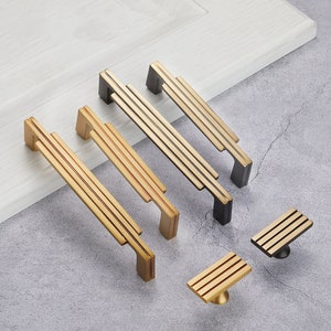 May include: A set of five gold and black cabinet door pulls with a geometric design. The pulls are made of metal and have a brushed finish.