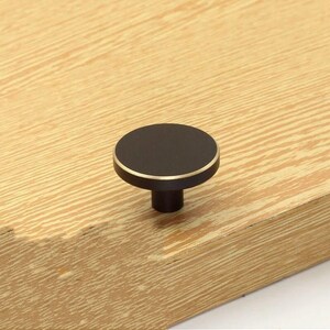May include: Black round cabinet knob with a gold trim. The knob is mounted on a wooden surface.