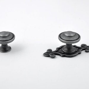 Może przedstawiać: Dwa antyczne srebrne gałki do szafek. Jeden to okrągła gałka z ozdobnym pierścieniem wokół górnej części. Drugi to prostokątna gałka z ozdobnym pierścieniem wokół górnej części i ozdobnym wzorem w postaci zwijanego ornamentu po obu stronach.