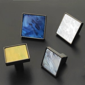 Peut inclure: Quatre boutons de cabinet carrés avec un cadre en métal noir et des inserts de différentes couleurs. Les inserts sont bleus, blancs, jaunes et gris.