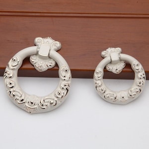 May include: Two white and gold decorative cabinet door pulls with ornate designs. The pulls are round with a ring handle.