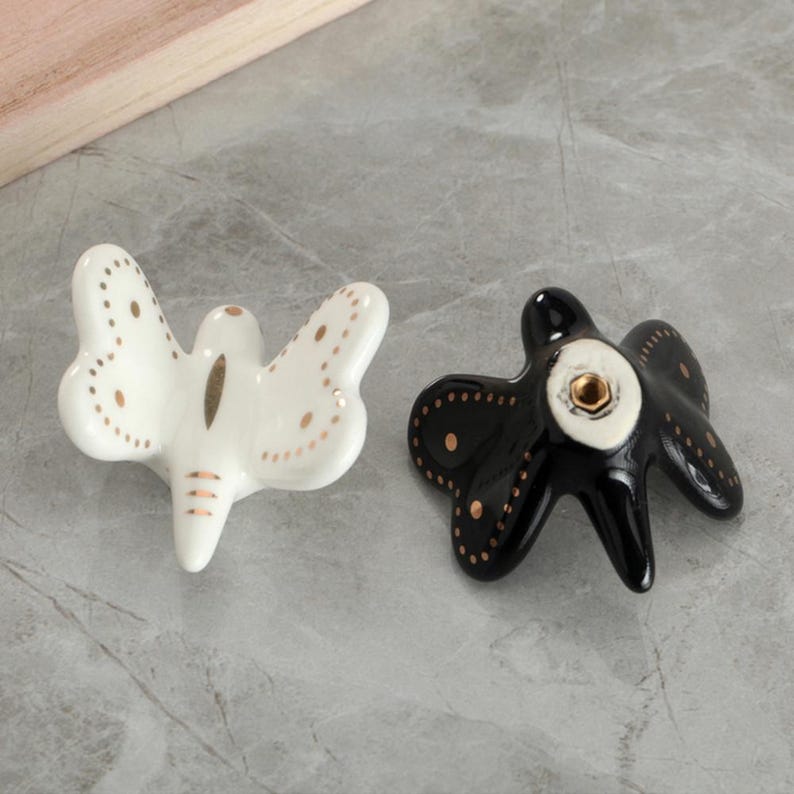 May include: Two ceramic butterfly-shaped cabinet knobs. One is white with gold-colored accents, and the other is black with gold-colored accents. The knobs are designed for furniture and have a decorative aesthetic.