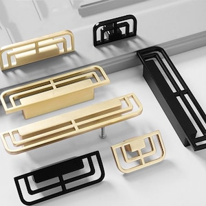 Peut inclure: Un ensemble de six poignées de meuble modernes en finitions noire et dorée. Les poignées présentent un design géométrique avec une forme rectangulaire et une série de carrés concentriques.