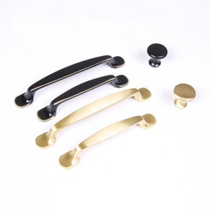 May include: A set of cabinet hardware featuring three black and two gold drawer pulls and two knobs. The pulls are curved and have a simple design. The knobs are round and have a smooth finish.