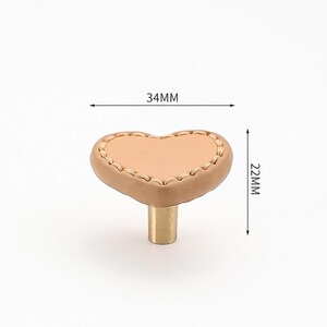 May include: A heart-shaped cabinet knob with a light brown leather surface and gold stitching. The knob measures 34mm wide and 22mm high.