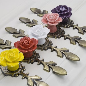 May include: Five antique-style cabinet knobs with ornate metal bases and ceramic rose flower accents. The roses are in various colors, including yellow, red, white, pink, and purple.