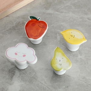 May include: Four ceramic cabinet knobs with fruit and cloud designs. The knobs include a red apple, a yellow lemon, a white cloud with pink trim, and a green pear, each with a smiling face. The knobs have a white, rounded base.