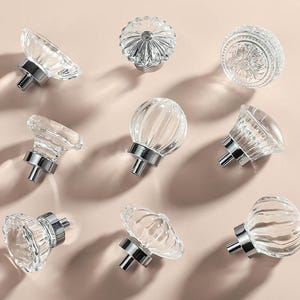May include: Assortment of clear glass cabinet knobs with silver metal bases. The knobs have various shapes and textures, including round, fluted, and patterned.