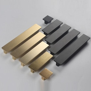 Pode incluir: Um conjunto de puxadores de armário em ouro escovado e preto em vários tamanhos. Os puxadores são de forma retangular com bordas arredondadas.