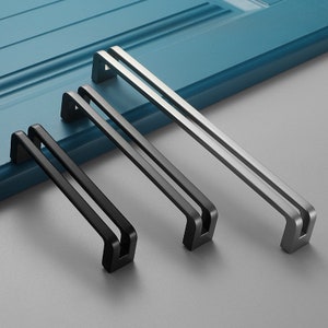 May include: Three cabinet pulls in different sizes. The largest pull is silver, the middle pull is black, and the smallest pull is black. All three pulls have a rectangular shape with a double-bar design.