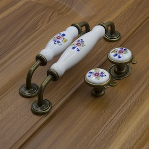 May include: Two white ceramic cabinet knobs with floral designs and antique brass hardware. The knobs are shown on a wood surface.
