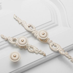 May include: Two white cabinet knobs with ornate gold accents and a floral design. The knobs are round and have a raised center.