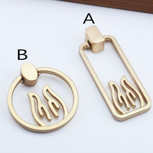 May include: Two gold-toned cabinet knobs with a flame design. The knob on the left is round with a ring handle. The knob on the right is rectangular with a straight handle.