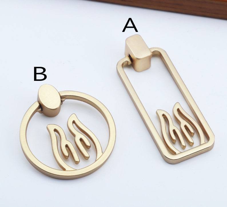 May include: Two gold-toned cabinet knobs with a flame design. The knob on the left is round with a ring handle. The knob on the right is rectangular with a straight handle.