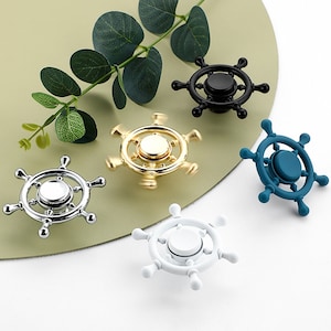 May include: Five different colored fidget spinners shaped like a ship's wheel. The spinners are gold, silver, black, white, and blue.