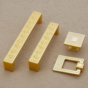 May include: Four gold cabinet knobs with a geometric pattern. Two are long and rectangular, one is square, and one is a pull with a square frame.