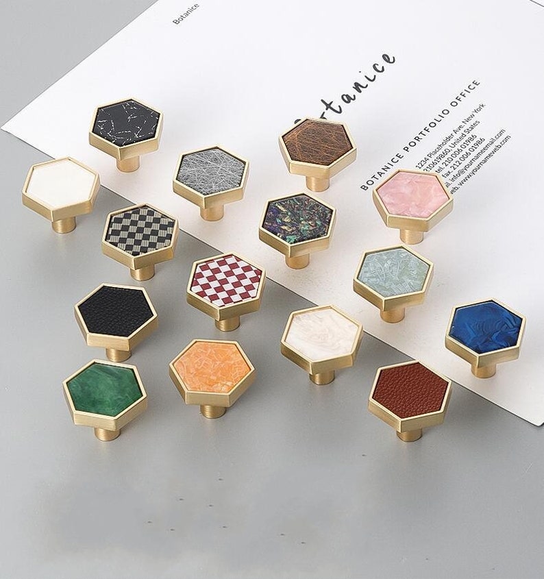 Pu&ograve; includere: Assortimento di 16 pomelli per armadi esagonali con vari motivi e colori. I pomelli sono in ottone e hanno una finitura dorata. I motivi includono marmo, pelle, a scacchi e disegni astratti.