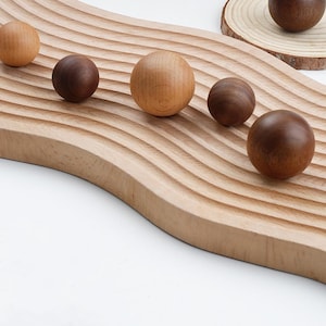 May include: A wooden tray with a wavy design features five wooden balls of different sizes and colours. The balls are arranged in a line across the tray.