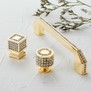 Puede incluir: Tres pomos de armario dorados con un diseño de cristal similar al diamante. Los pomos son de diferentes formas, incluyendo un cuadrado, un redondo y una manija curva.