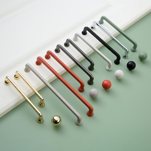 May include: Assortment of cabinet hardware pulls in various colors, including gold, white, gray, black, red, and green. The pulls are made of metal and have a rounded knob at the end.