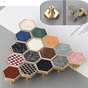 Könnte beinhalten: Ein Satz von 12 sechseckigen Schrankknöpfen mit verschiedenen Mustern. Die Knöpfe sind aus Messing gefertigt und haben eine goldene Oberfläche. Die Muster umfassen Marmor, Leder, Holz und karierte Designs. Die Knöpfe sind in einem Wabenmuster angeordnet.