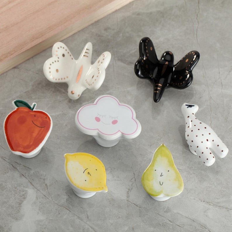 May include: A collection of ceramic cabinet knobs in various shapes and colors. Designs include a white butterfly with gold accents, a black dragonfly, a red apple, a white cloud with pink accents, a yellow lemon, a green pear, and a white bird with red dots.
