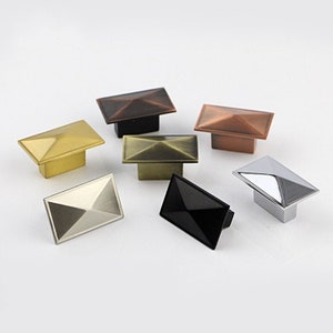 Op de afbeelding: Zes verschillende gekleurde kastknoppen met een piramidevorm. De knoppen zijn gemaakt van metaal en hebben een gepolijste afwerking. De kleuren zijn goud, koper, brons, zilver, zwart en antiek koper.