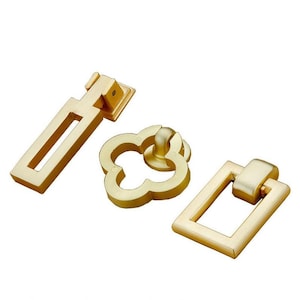 Può includere: Tre pomelli per armadi dorati in stili diversi. Uno è un semplice quadrato con un tirare ad anello, un altro è un design a quadrifoglio con un pomo rotondo, e l'ultimo è un tirare rettangolare con un'apertura lunga e sottile.