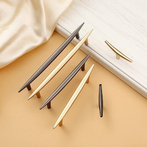 May include: A selection of contemporary cabinet handles in gold, bronze, and black. The handles have a streamlined, tapered design. Some are long and slender, while others are T-shaped. Measurements are in centimetres.