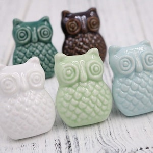 May include: Five ceramic owl drawer pulls in different colours: white, green, blue, brown, and teal. Each owl has a detailed design with raised eyes and feathers.