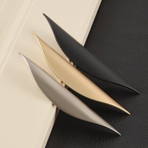 May include: Three cabinet pulls in different finishes: black, gold, and silver. The pulls are shaped like a leaf and have a curved design.